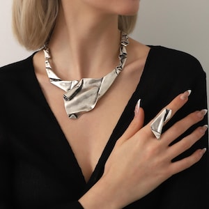 May include: A silver-toned necklace and ring set. The necklace has a unique, abstract design with folded metal elements. The ring mirrors the necklace's style. The jewelry is worn against a black top, creating a striking contrast.