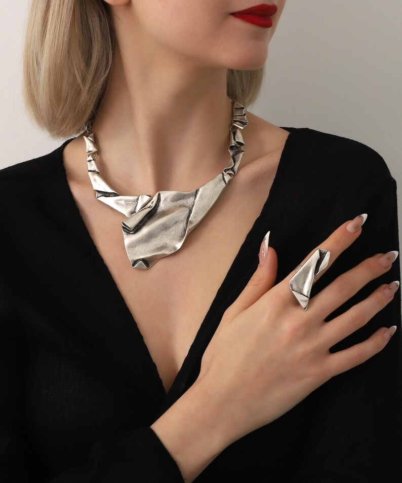May include: A silver-toned necklace and ring set. The necklace has a unique, abstract design with folded metal elements. The ring mirrors the necklace's style. The jewelry is worn against a black top, creating a striking contrast.