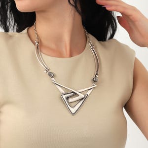 May include: A silver necklace with a geometric pendant featuring a layered triangle design. The pendant has a textured surface and is accented with small silver beads.