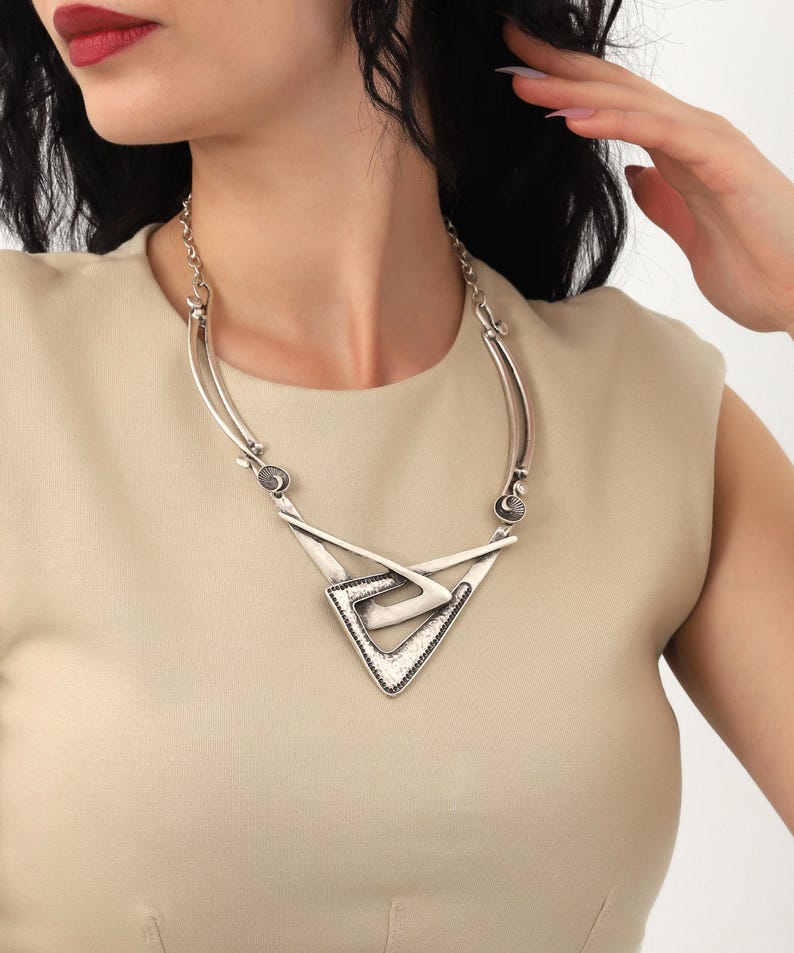 May include: A silver necklace with a geometric pendant featuring a layered triangle design. The pendant has a textured surface and is accented with small silver beads.
