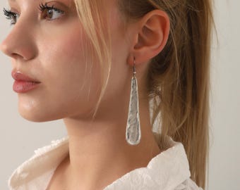 May include: A long, teardrop-shaped earring with a textured, hammered silver finish. The earring features a smooth, rounded bottom edge and tapers toward the top, where it connects to a thin, dark metal hook for attachment to the earlobe.