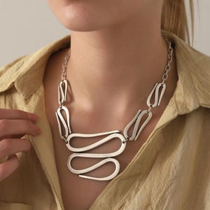 May include: A silver-toned necklace featuring a series of interconnected, elongated, and curved metal links. The design consists of multiple smooth, abstract, wave-like shapes that form a central focal point, suspended from a simple metal chain.