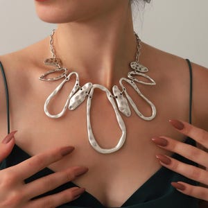 May include: A silver-toned statement necklace with an abstract design. The necklace features irregular, textured shapes and chain links. The jewelry is worn against a dark green top. The model's hands are visible, with matching nail polish.