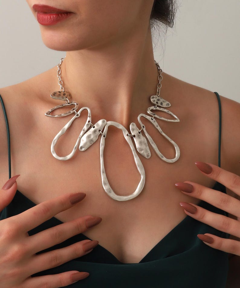 May include: A silver-toned statement necklace with an abstract design. The necklace features irregular, textured shapes and chain links. The jewelry is worn against a dark green top. The model's hands are visible, with matching nail polish.