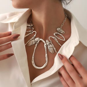 May include: Silver-toned statement necklace with an abstract, hammered metal design. The necklace features a large, teardrop-shaped pendant and various organic shapes connected by a delicate chain. The jewelry is worn over a white collared shirt.