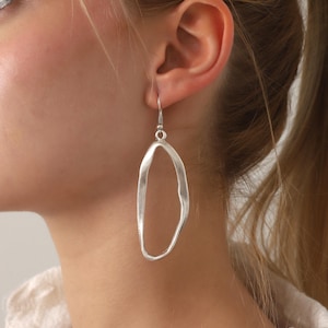 May include: A silver-toned dangle earring featuring an elongated, irregular, and organic open-loop shape with a matte, textured finish. The earring hangs from a simple silver-toned hook wire, creating a long, slender silhouette that rests against the neck.