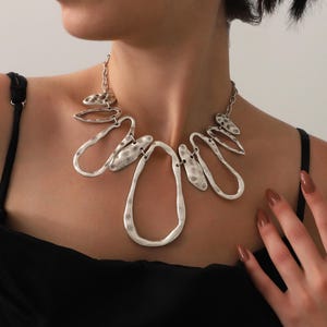 May include: A silver-toned statement necklace with an abstract design. The necklace features hammered metal shapes, including teardrops and ovals, connected by a chain. The jewelry is worn against a black top.