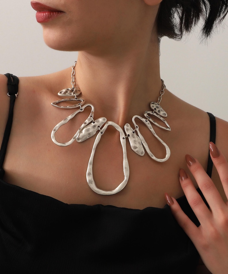 May include: A silver-toned statement necklace with an abstract design. The necklace features hammered metal shapes, including teardrops and ovals, connected by a chain. The jewelry is worn against a black top.