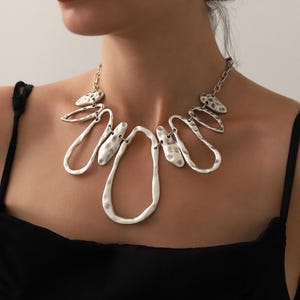 May include: Silver-toned statement necklace with an abstract design. The necklace features a series of interconnected, textured metal shapes, including elongated teardrops and irregular ovals. The necklace is worn against a black top.