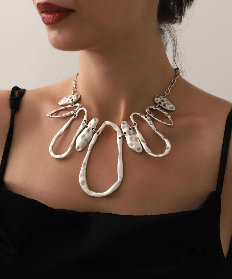 May include: Silver-toned statement necklace with an abstract design. The necklace features a series of interconnected, textured metal shapes, including elongated teardrops and irregular ovals. The necklace is worn against a black top.