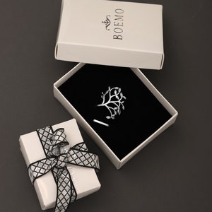 May include: A silver ring with a tree design in a white box with black interior. The box has the word "BOEMO" printed on the top.