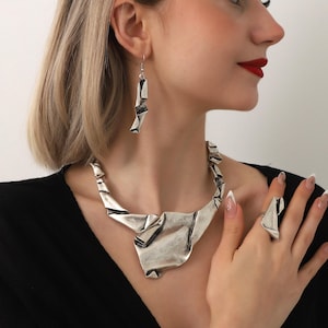 May include: A silver-toned jewelry set featuring a necklace, earrings, and a ring. The necklace and ring have a folded, abstract design. The earrings are long and dangle. The jewelry is worn against a black top.