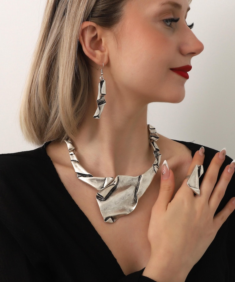 May include: A silver-toned jewelry set featuring a necklace, earrings, and a ring. The necklace and ring have a folded, abstract design. The earrings are long and dangle. The jewelry is worn against a black top.