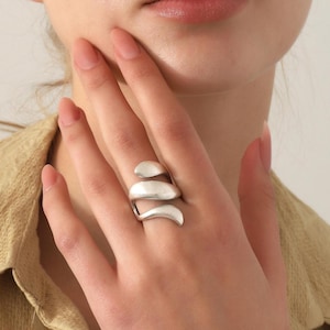 Peut inclure: Une bague argentée au design croisé composée de trois bandes lisses, incurvées et superposées qui s'enroulent autour du doigt. Le métal présente une finition mate et brossée, offrant un éclat doux sur ces formes organiques fluides.