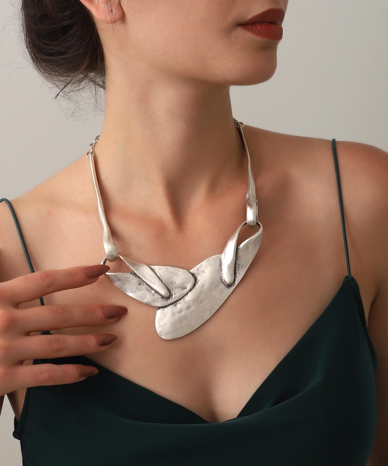 May include: Silver-toned statement necklace with an abstract design. The necklace features a hammered texture and a unique shape, with a large central pendant and delicate chain. The jewelry is worn against a dark green top.