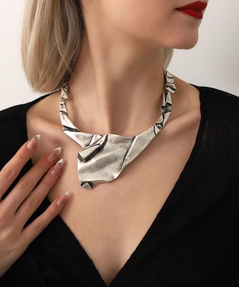 May include: A silver metal necklace with a unique, abstract design. The necklace is made of a single piece of metal that is folded and twisted into a shape that resembles a crumpled piece of paper. The necklace is a statement piece that would add a touch of modern style to any outfit.