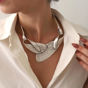 May include: A silver-toned statement necklace with an abstract design. The necklace features a large, textured pendant with curved and angular elements. The jewelry is worn over a white collared shirt.