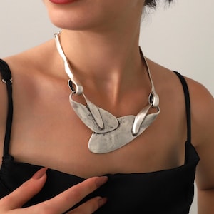 May include: A silver-toned statement necklace with an abstract design. The necklace features a series of interconnected, textured metal elements, including elongated curved pieces and a central, leaf-like shape. The model is wearing a black top.