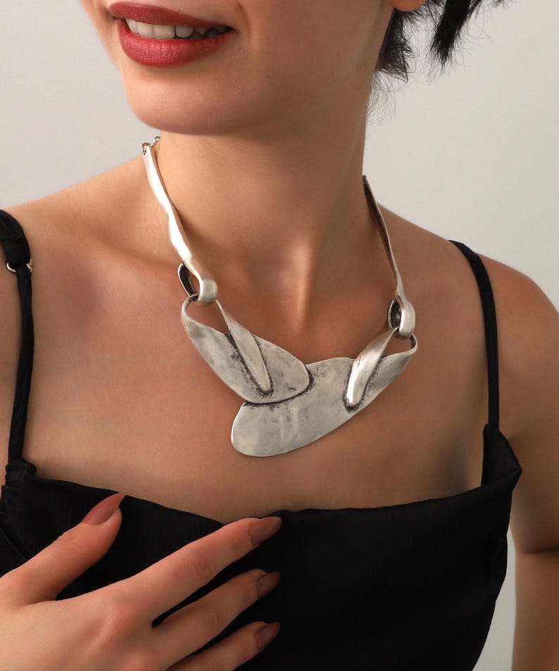 May include: A silver-toned statement necklace with an abstract design. The necklace features a series of interconnected, textured metal elements, including elongated curved pieces and a central, leaf-like shape. The model is wearing a black top.
