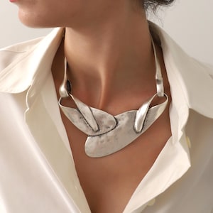 May include: A silver-toned statement necklace with an abstract design. The necklace features a large, textured central element and curved, elongated pieces that connect to the chain. The jewelry is worn over a white collared shirt.