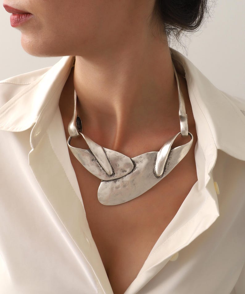 May include: A silver-toned statement necklace with an abstract design. The necklace features a large, textured central element and curved, elongated pieces that connect to the chain. The jewelry is worn over a white collared shirt.