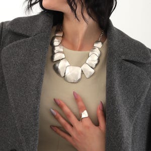 May include: A silver necklace with large, irregular, flat, geometric links. The necklace is worn by a person with dark hair and a gray coat. The person is also wearing a silver ring on their finger.