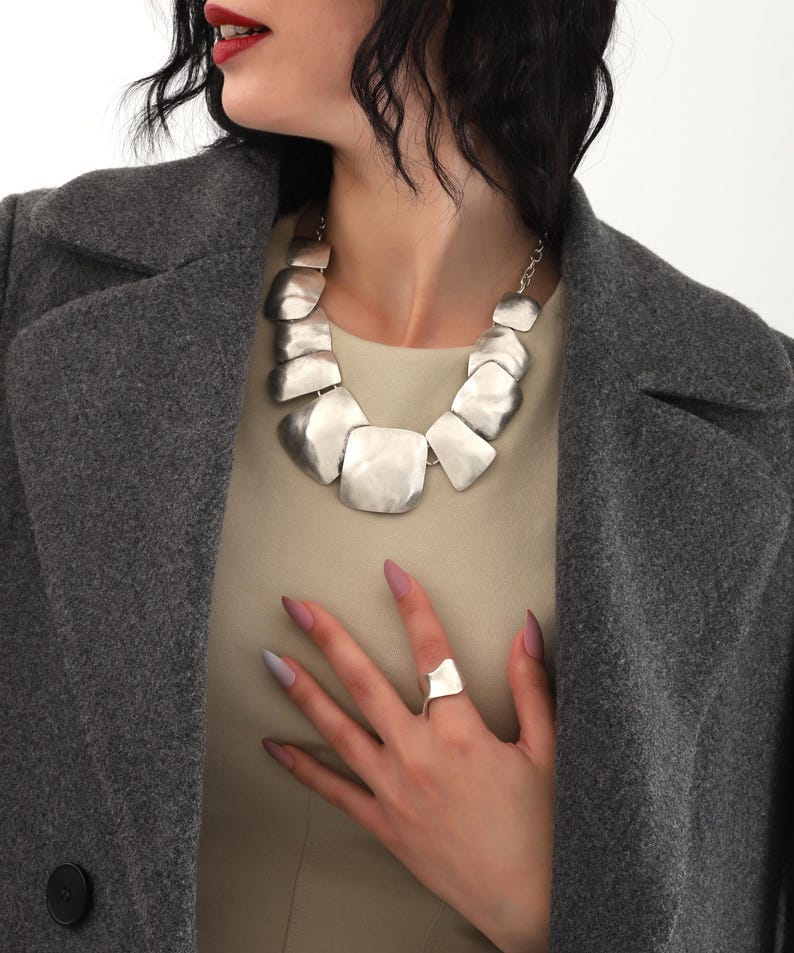 May include: A silver necklace with large, irregular, flat, geometric links. The necklace is worn by a person with dark hair and a gray coat. The person is also wearing a silver ring on their finger.