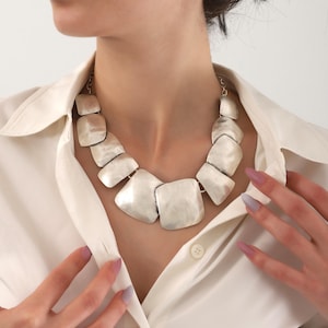 May include: A silver statement necklace with large, flat, square-shaped links. The necklace is worn by a person with a white shirt.