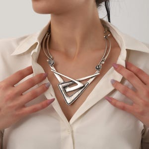 May include: A silver statement necklace with a geometric design featuring two overlapping triangles. The necklace has a textured finish and a silver chain.