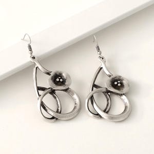 May include: A pair of silver-toned metal dangle earrings featuring an abstract, curved frame design with a central flower-like element containing three small raised dots. The earrings have a dark, oxidized finish and include standard hook-style wires.
