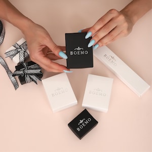 May include: An overhead shot shows several small gift boxes in black and white, some with decorative bows. The boxes are labeled "BOEMO" in a stylized font. The scene is set on a light pink surface, with hands holding a black box.