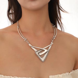 May include: A silver statement necklace with a geometric design. The necklace features a large, triangular pendant with a smaller, overlapping triangle inside. The pendant has a textured, metallic finish.