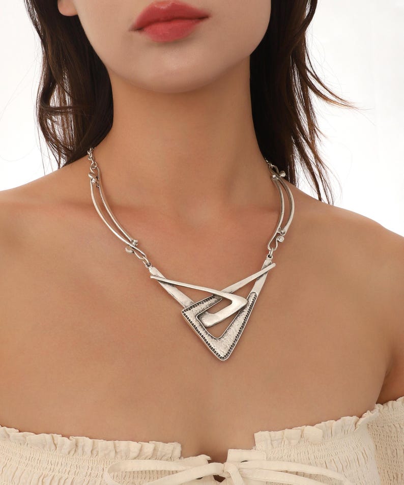 May include: A silver statement necklace with a geometric design. The necklace features a large, triangular pendant with a smaller, overlapping triangle inside. The pendant has a textured, metallic finish.