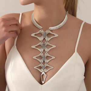 May include: A silver-toned statement necklace featuring a rigid collar band connected to a vertical series of four diamond-shaped, textured metal links that taper downward, ending in a smaller, singular pointed metal pendant piece.