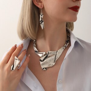 May include: A silver-toned jewelry set featuring a necklace, earrings, and ring. The necklace has a wide, textured design, while the earrings are long and angular. The ring has a matching geometric shape. The jewelry is worn with a white button-down shirt.