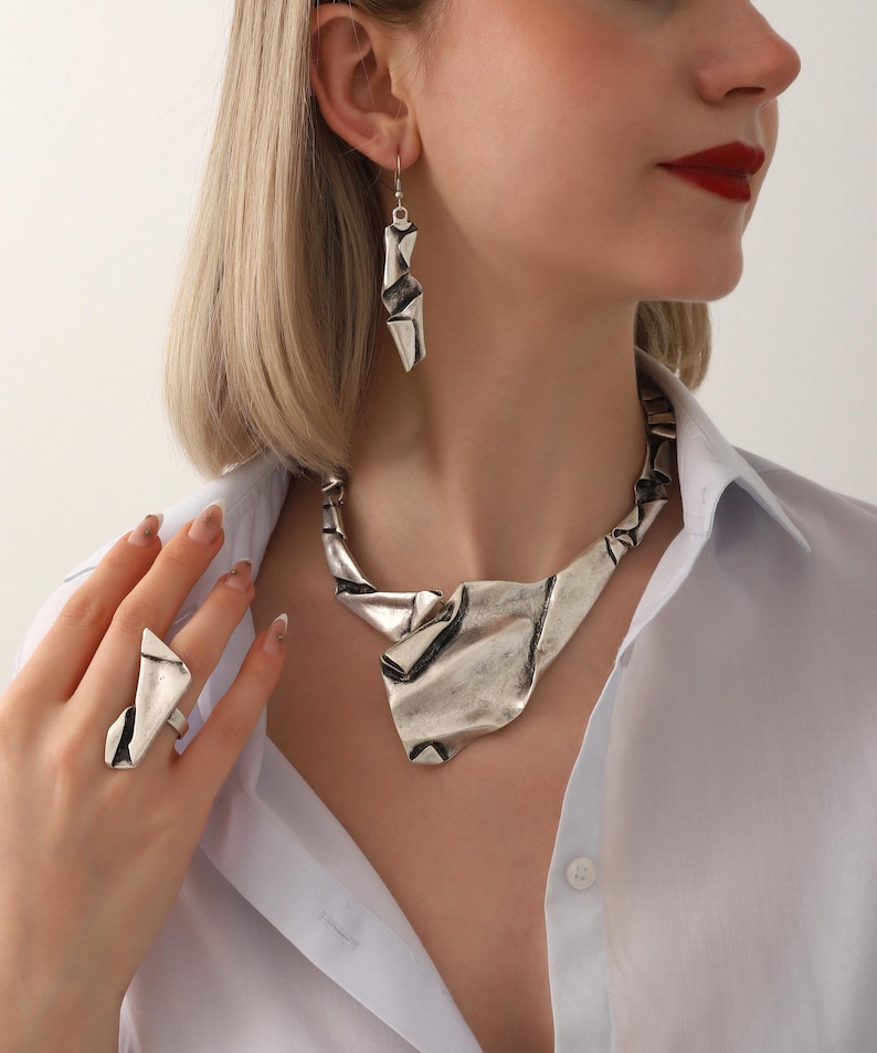 May include: A silver-toned jewelry set featuring a necklace, earrings, and ring. The necklace has a wide, textured design, while the earrings are long and angular. The ring has a matching geometric shape. The jewelry is worn with a white button-down shirt.