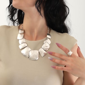 May include: A silver statement necklace with large, flat, geometric links. The necklace is worn by a person with long, dark hair and nude nail polish. The person is also wearing a silver ring on their finger.