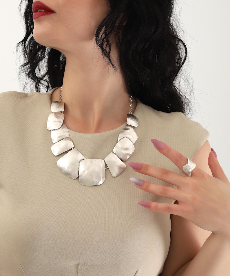 May include: A silver statement necklace with large, flat, geometric links. The necklace is worn by a person with long, dark hair and nude nail polish. The person is also wearing a silver ring on their finger.