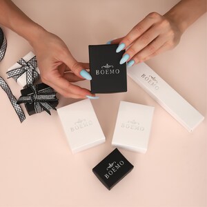 May include: Five small boxes with the word "BOEMO" printed on them. The boxes are white and black and are arranged on a light pink surface.