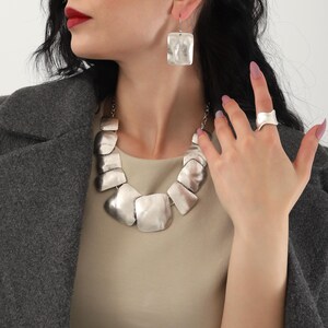 May include: A silver necklace with large, irregular, flat, silver links. The necklace is paired with matching silver earrings and a silver ring.