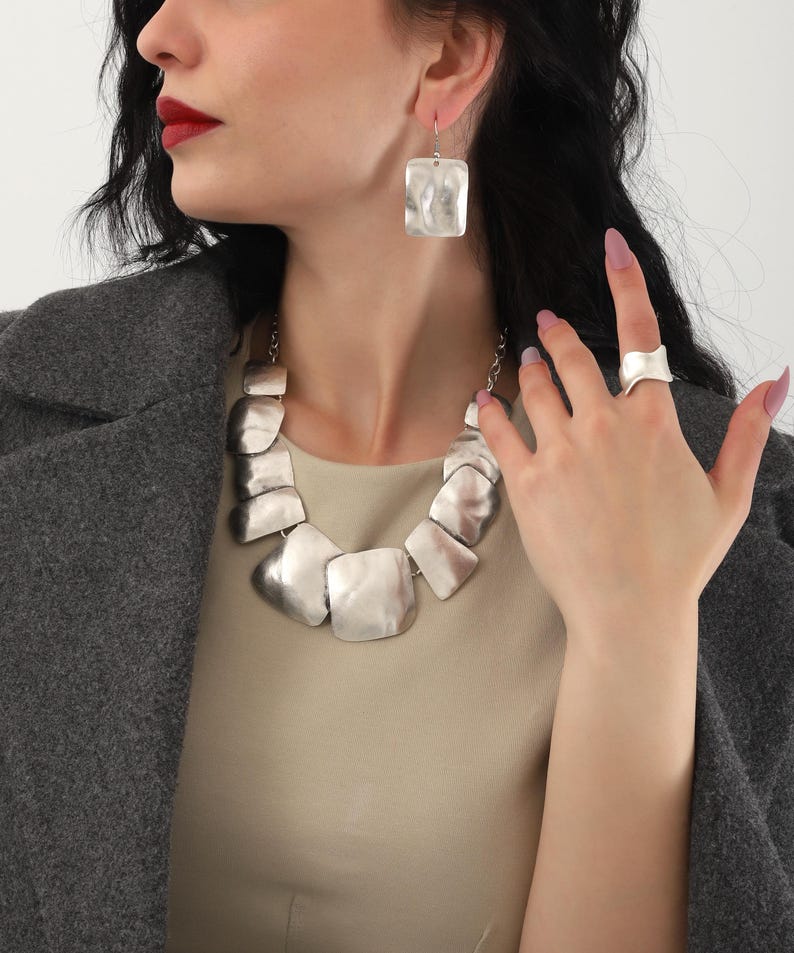 May include: A silver necklace with large, irregular, flat, silver links. The necklace is paired with matching silver earrings and a silver ring.