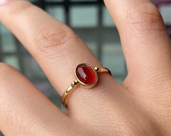 May include: A gold ring featuring an oval, reddish-orange gemstone. The ring has a thin band and small decorative beads on each side of the gemstone. The ring is shown on a person's finger.