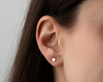 May include: A close-up of a person's ear with a small, square-shaped gold earring.