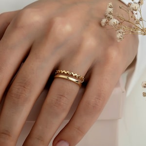 May include: Two gold rings on a finger. The top ring is a delicate chain of small, round beads. The bottom ring is a simple, flat band.