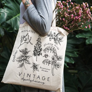 May include: A white canvas tote bag with black illustrations of poisonous herbs, including mistletoe, henbane, bleeding heart, and foxglove. The text "Vintage Apothecary" is printed on the bag.
