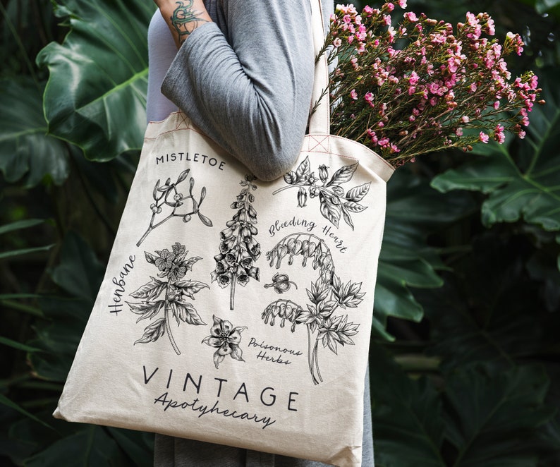 May include: A white canvas tote bag with black illustrations of poisonous herbs, including mistletoe, henbane, bleeding heart, and foxglove. The text "Vintage Apothecary" is printed on the bag.