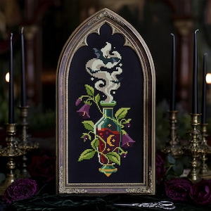 May include: A cross-stitch embroidery in a pointed arch frame featuring a glass bottle filled with red liquid, a yellow crescent moon, green leaves, and purple bell-shaped flowers, with white smoke rising from the bottle top.