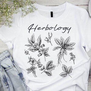 May include: White t-shirt with the word "Herbology" printed in black. The shirt features a black and white illustration of various herbs and plants.