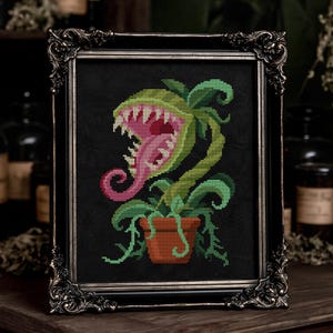 May include: A cross-stitch embroidery of a green carnivorous plant with a pink interior and sharp white teeth, potted in a brown container. The artwork is displayed within an ornate, dark metallic frame with intricate scrollwork details.