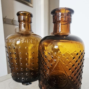 Könnte beinhalten: Zwei bernsteinfarbene Glasflaschen mit engem Hals und breitem Rand. Die linke Flasche hat eine genoppte Textur und den geprägten Text TO BE TAKEN. Die rechte Flasche weist ein Rautenmuster und eine dreieckige Prägung auf.